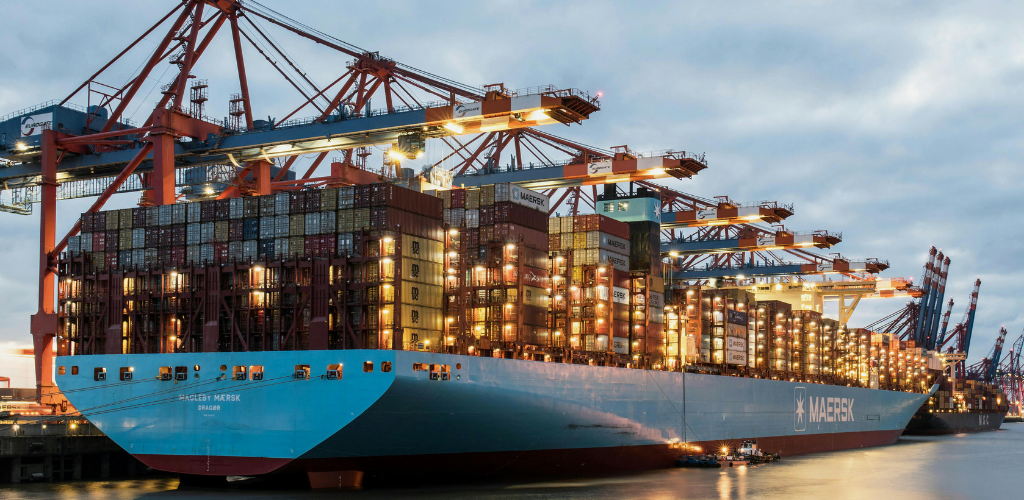 A large Maersk container ship docked at a commercial port during twilight, being loaded by massive orange gantry cranes under industrial lighting. A large Maersk container ship docked at a commercial port during twilight, being loaded by massive orange gantry cranes under industrial lighting.